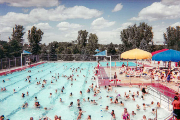 Outdoor public swimming pool with many people in the water and on the deck. Trees and a fence are in the background under a partly cloudy sky.