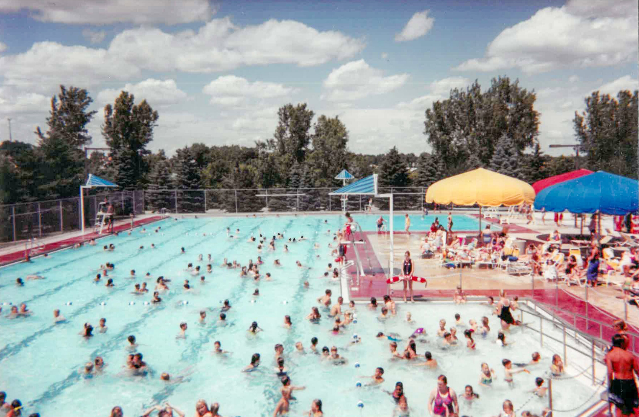 Outdoor public swimming pool with many people in the water and on the deck. Trees and a fence are in the background under a partly cloudy sky.