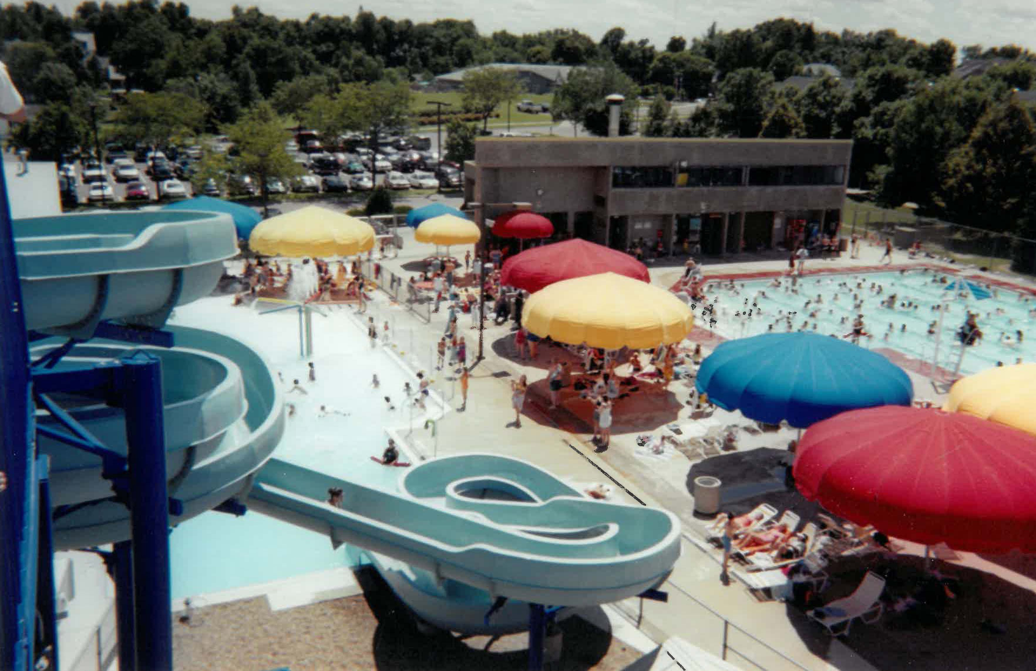 A busy outdoor public pool with waterslides, colorful umbrellas, and many people swimming and lounging on a sunny day.