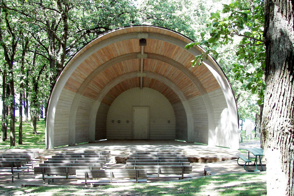An empty outdoor bandshell with wooden benches in front, surrounded by trees in a park setting.