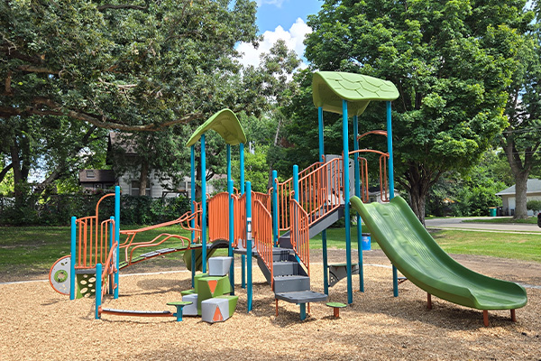 Outdoor playground with green and orange slides, climbing structures, and steps on a mulch surface, surrounded by trees and grass.