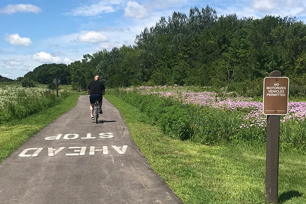 A person rides a bicycle on a paved trail through a green landscape with wildflowers. A sign reads 