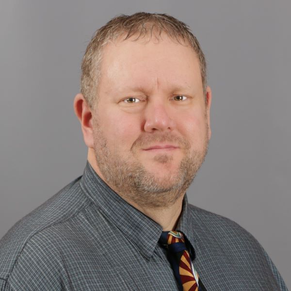 Portrait of a man with short brown hair, wearing a gray patterned shirt and a colorful tie, against a gray background