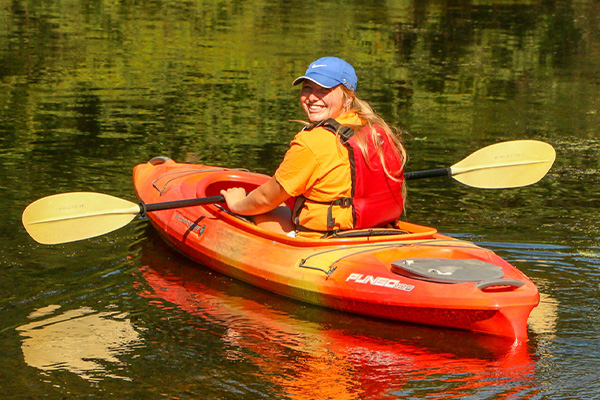 Person wearing an orange shirt, red life vest, and blue cap paddling a red kayak on calm water, looking back and smiling.
