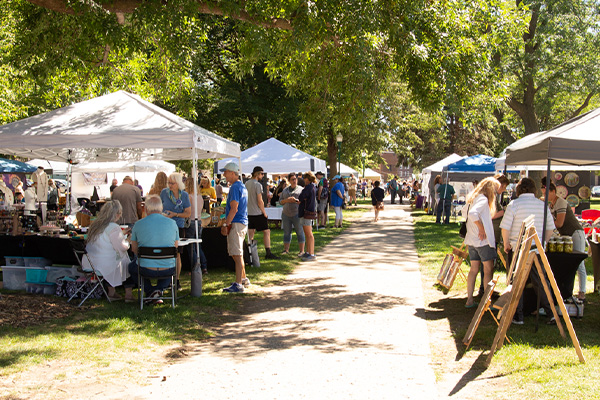 People browse booths and interact with vendors at an outdoor market under white tents on a sunny day in a park.