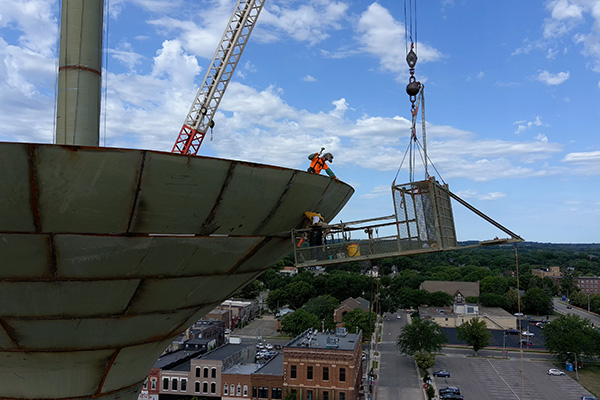 Two workers in safety gear stand on the edge of a large, elevated metal structure while a crane lifts a metal framework beside them above a town.
