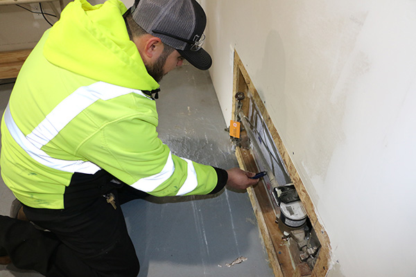 A person in a reflective jacket and cap kneels on the floor, using a tool to work on exposed pipes in a wall cutout.
