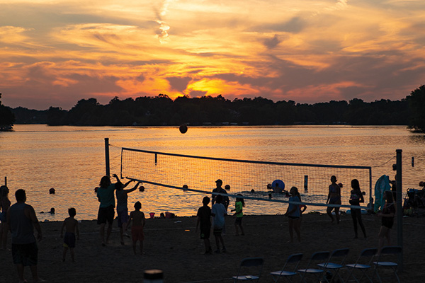People play volleyball on a sandy beach beside a lake at sunset, with silhouettes visible against the colorful sky and water.