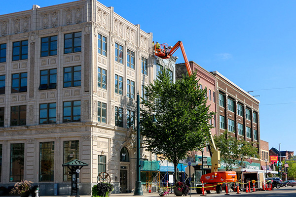 Cherry picker lifting workers to a building facade in an urban setting, surrounded by trees and adjacent buildings under a clear blue sky.