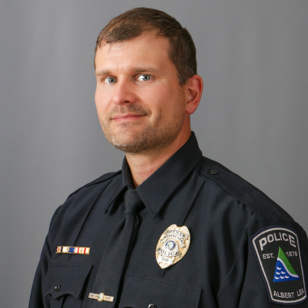 Dan Schmitt, A police officer in uniform stands against a gray background, displaying badges on his shirt and shoulder.