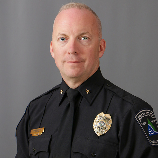 Darren Hanson, A man in a police uniform stands in front of a gray background, wearing a badge and insignia on his shirt.