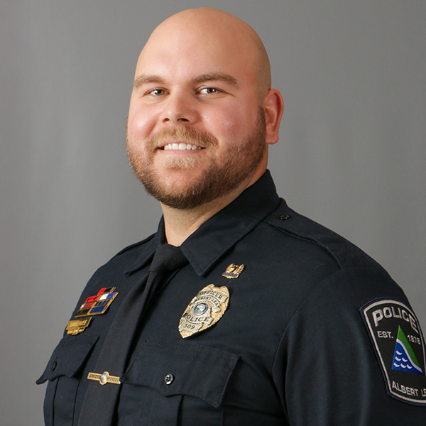 David Huse, A man in a police uniform smiles against a gray background, displaying badges on his shirt.