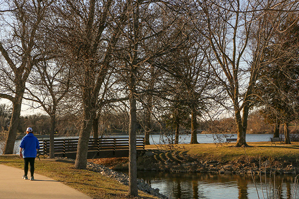 A person wearing a blue jacket walks on a paved path near a leafless tree-lined river, with a small bridge and grassy area in the background.