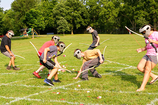 Children wearing protective masks play archery tag on a grassy field, holding bows and collecting foam-tipped arrows. Trees and a playground are visible in the background.