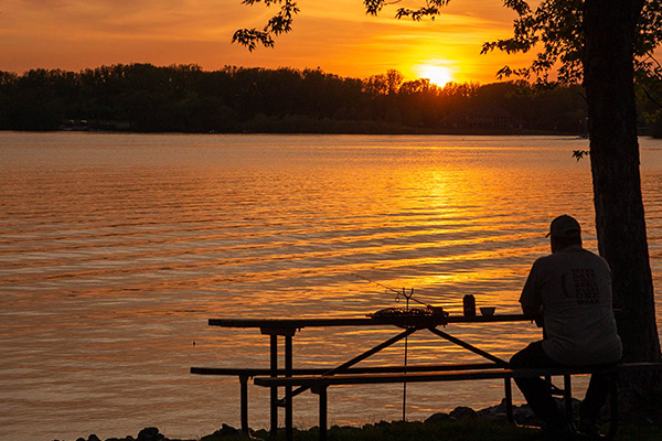 A person sits at a picnic table by a lake at sunset, with trees silhouetted in the background and orange sunlight reflecting on the water.