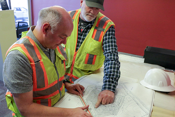 Two men in safety vests discuss a construction blueprint on a table indoors. A hard hat is placed nearby.