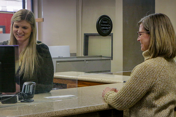 A woman stands at a bank counter, speaking with a teller behind a glass partition. The teller is seated with a computer in front of her.