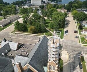 Aerial view of a church with a scaffolding-covered tower, nearby streets, parked cars, trees, and buildings in the background.