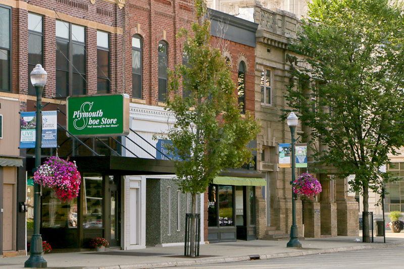 Brick storefronts line a small-town street with trees, hanging flower baskets, and a green sign for Plymouth Shoe Store visible in the foreground.