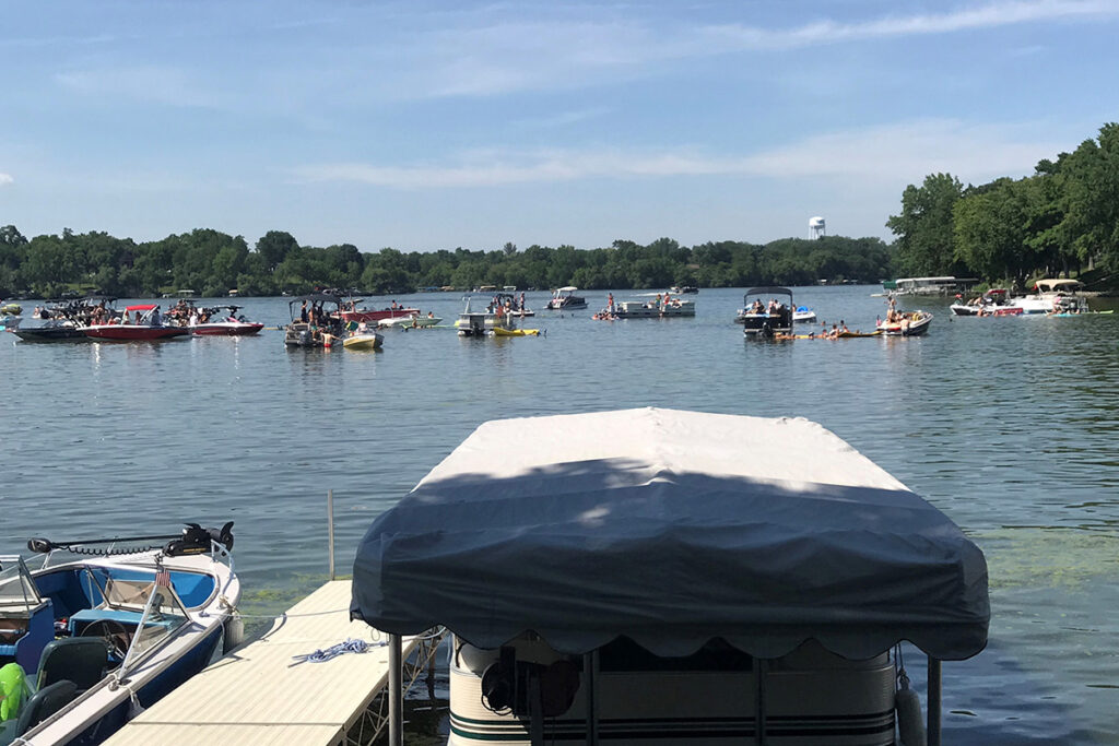 Boats and people on a lake on a sunny day. Trees and a water tower are visible on the shoreline.