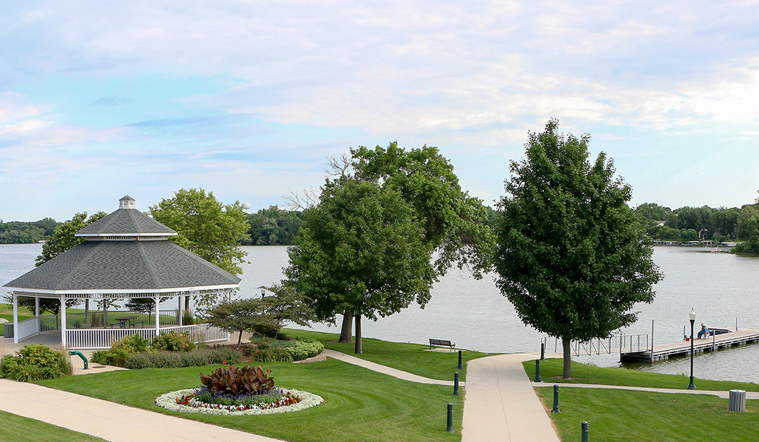 A gazebo stands near a flower bed and pathway by a lake, with trees, a dock, and benches visible under a partly cloudy sky.