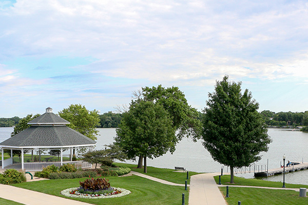 A riverside park with a white gazebo, landscaped flower bed, paved paths, green trees, and a dock extending into the water under a partly cloudy sky.