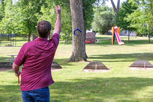 A man in a red striped shirt throws a horseshoe toward a stake in a grassy park with trees, picnic tables, and a playground in the background.