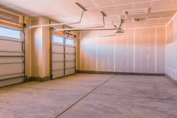Empty two-car garage with unfinished drywall, concrete floor, and two closed garage doors with windows at the top. Ceiling has exposed garage door mechanisms and attic access panel.