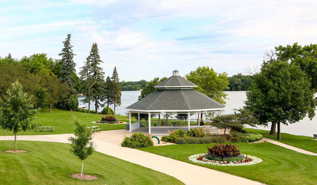 A white gazebo stands near a lake, surrounded by green lawns, trees, pathways, and flowerbeds under a partly cloudy sky.