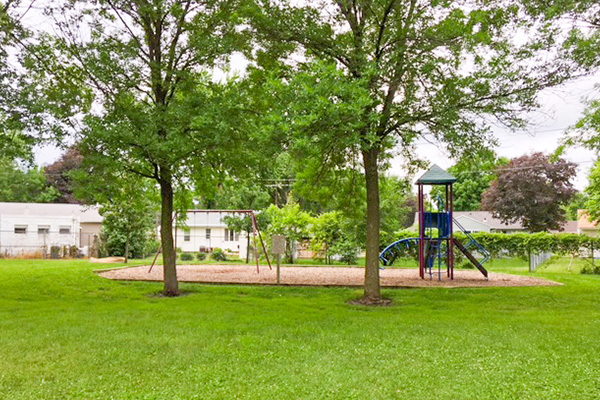 Playground with swings and a slide beneath large trees, surrounded by grass, with houses and trees in the background.
