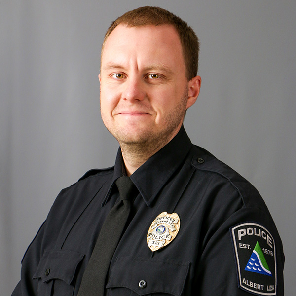 Gregg Calbick, A uniformed police officer stands against a neutral background, wearing a black shirt with a police badge and shoulder patch, looking straight ahead.