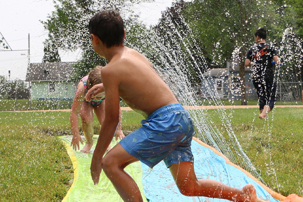 Children play on a slip and slide with water spraying around them on a grassy lawn, with houses and trees visible in the background.