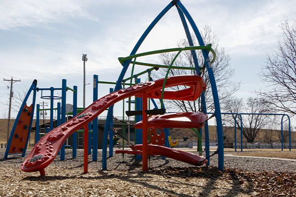 A playground structure with a red climbing slide and blue support beams stands on wood chips, with swings and leafless trees in the background.