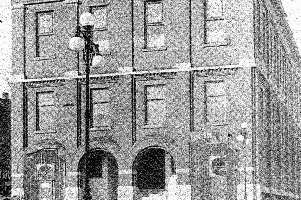 Historic black-and-white photo of a three-story brick building with arched doorways and a streetlamp in front. Handwritten text appears in the lower right corner.