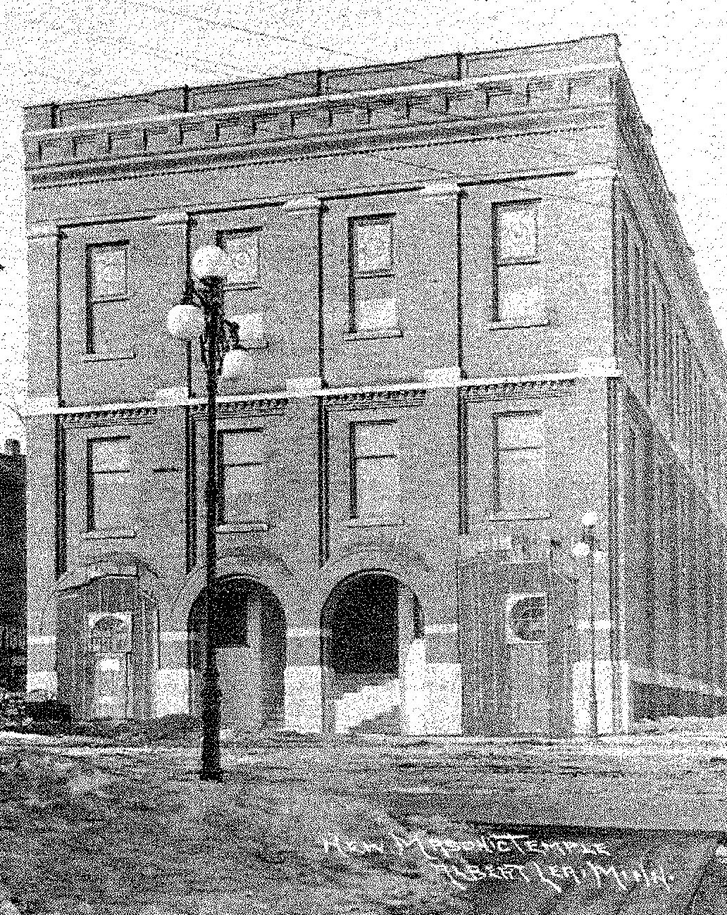 Historic black-and-white photo of a three-story brick building with arched doorways and a streetlamp in front. Handwritten text appears in the lower right corner.