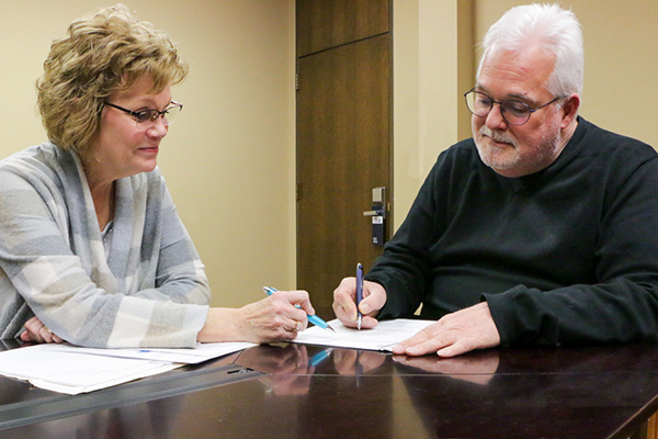 Two people sit at a table, reviewing and signing documents. They are focused on the papers, with pens in hand, suggesting a discussion or agreement.