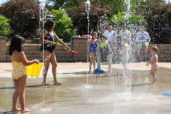 Children and an adult play in swimsuits at a splash pad with water jets spraying, while several adults sit on a bench in the background.
