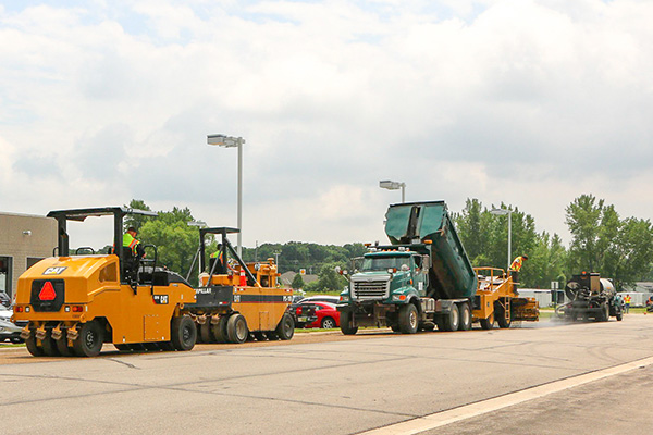 Road construction crew resurfacing a road using heavy machinery, including a dump truck unloading material and a steamroller compacting the surface.