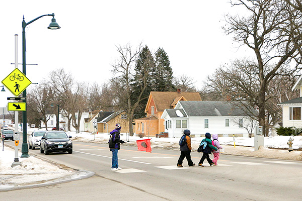 A crossing guard helps three children cross a snowy suburban street at a crosswalk on a cloudy day.
