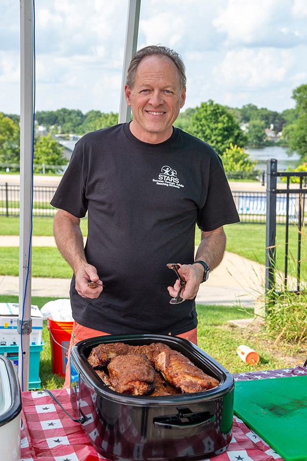 A man stands outdoors behind a table with a red checkered tablecloth, smiling and holding tongs over a slow cooker filled with seasoned meat.