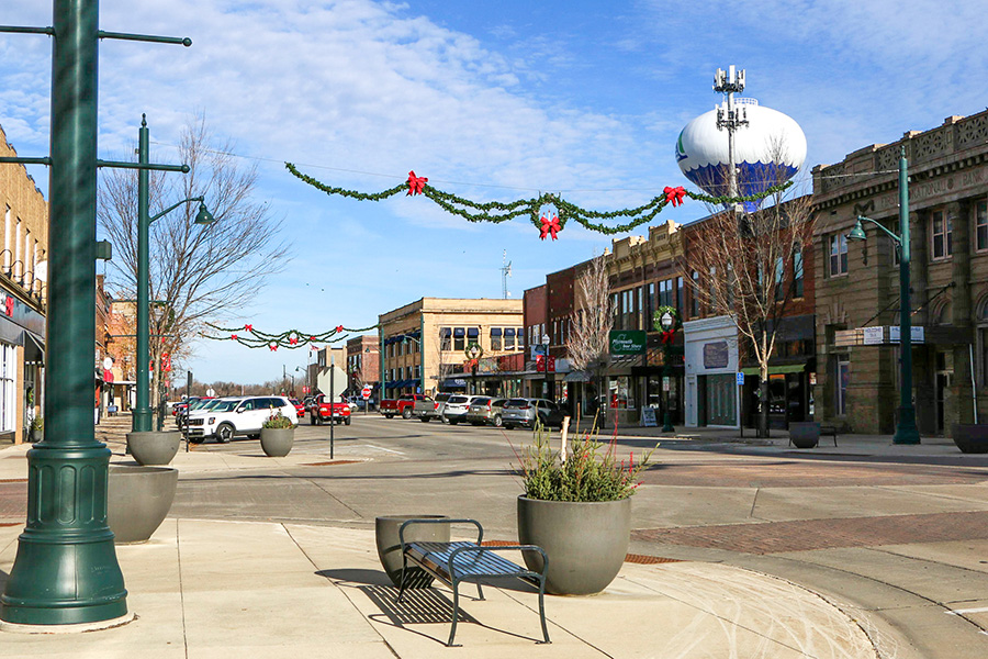 A small town main street with storefronts, parked cars, holiday garlands overhead, and a water tower in the background under a blue sky.