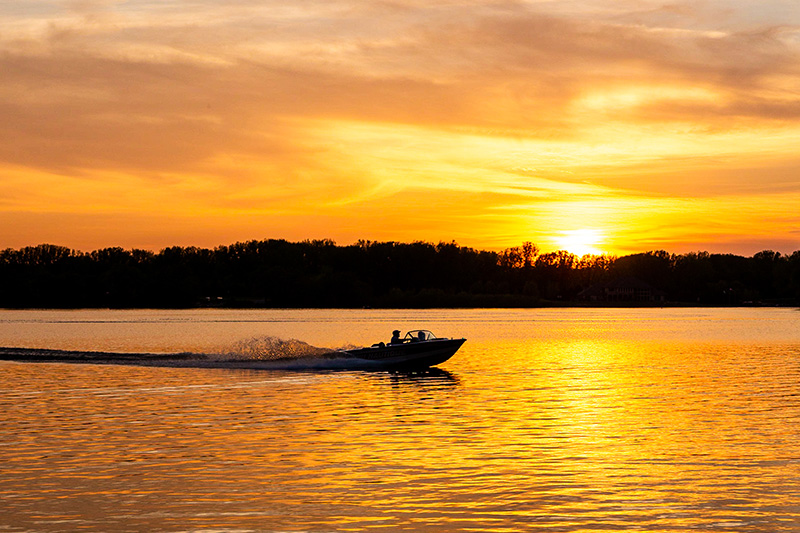 A motorboat speeds across a calm lake at sunset, with the sun setting behind a line of trees, casting orange and yellow hues over the water.