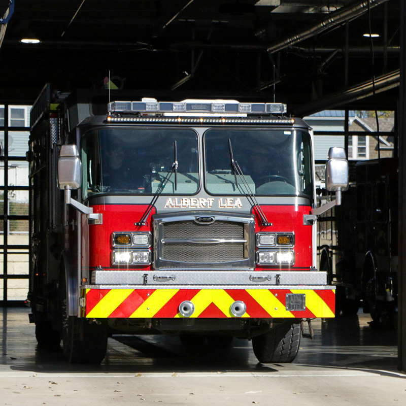 A red fire truck labeled "Albert Lea" is parked inside a fire station garage, with sunlight illuminating the front of the vehicle.