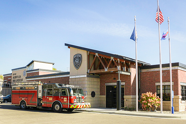 A red fire truck is parked in front of a brick fire station with three flagpoles displaying flags and a department emblem on the building.