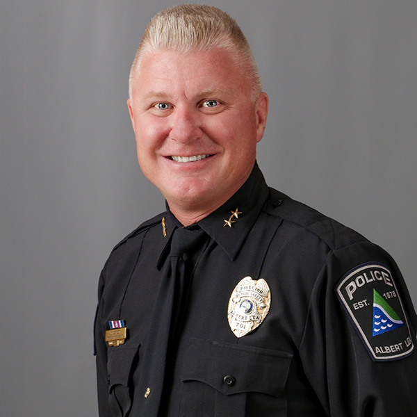 J.D. Carlson, A person in a police uniform, with short blond hair, smiles against a gray background. There are badges on the shirt and shoulder.