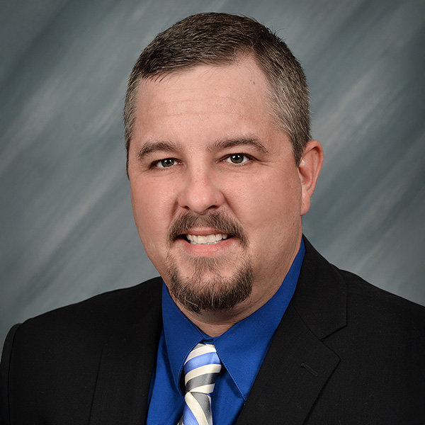 Man with short brown hair and goatee, wearing a dark suit, blue shirt, and striped tie, posed in front of a gray studio backdrop.
