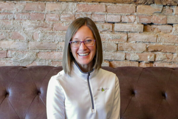 A woman with straight, shoulder-length hair and glasses sits on a brown sofa in front of a brick wall, smiling at the camera. She wears a light-colored, zip-up top.