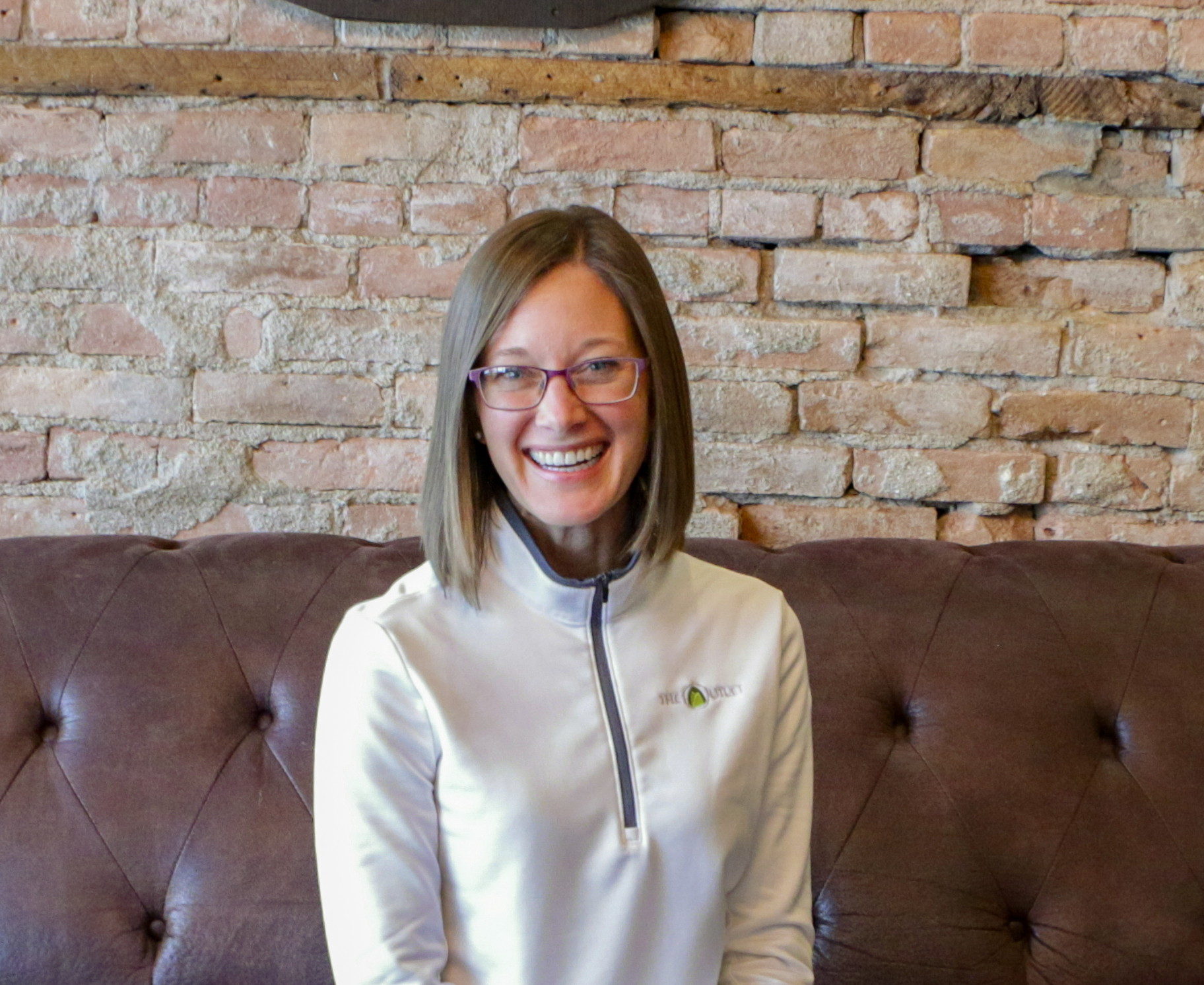 A woman with straight, shoulder-length hair and glasses sits on a brown sofa in front of a brick wall, smiling at the camera. She wears a light-colored, zip-up top.