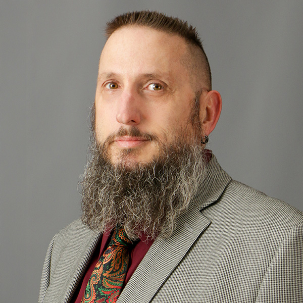 Keith Van Beek, A person with a long beard and earrings is wearing a gray blazer, patterned tie, and maroon shirt against a gray background.
