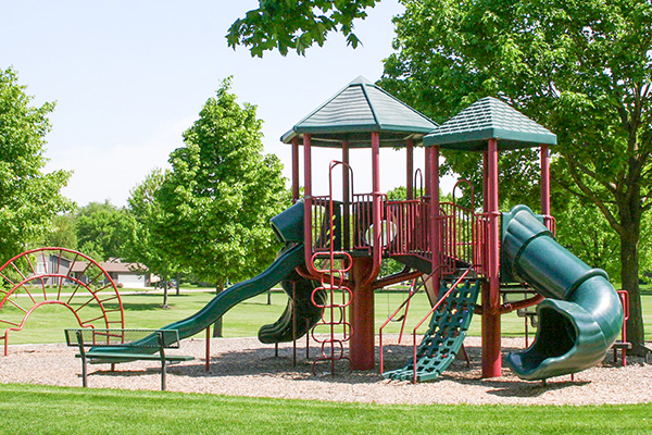A playground with slides, climbing structures, and benches sits on a grassy area surrounded by trees on a sunny day.